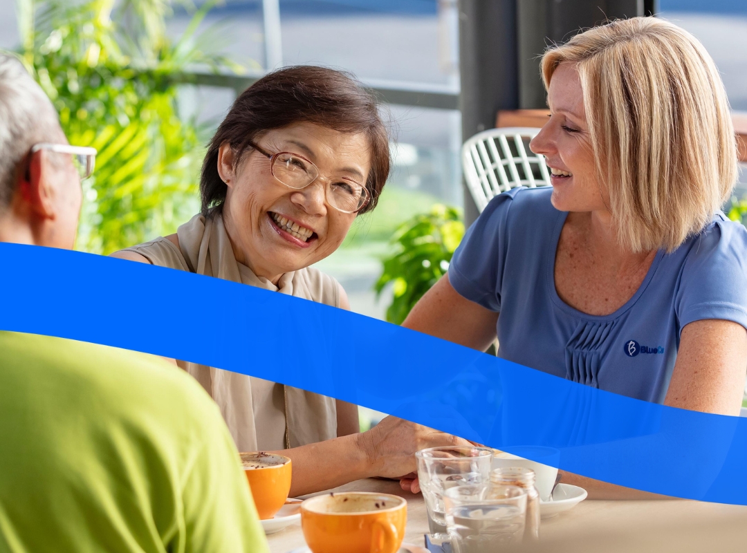 Three people sitting at a cafe table with coffee cups and glasses. One person is wearing a green shirt, another a beige scarf, and the third a blue top. They are all smiling and engaged in conversation.