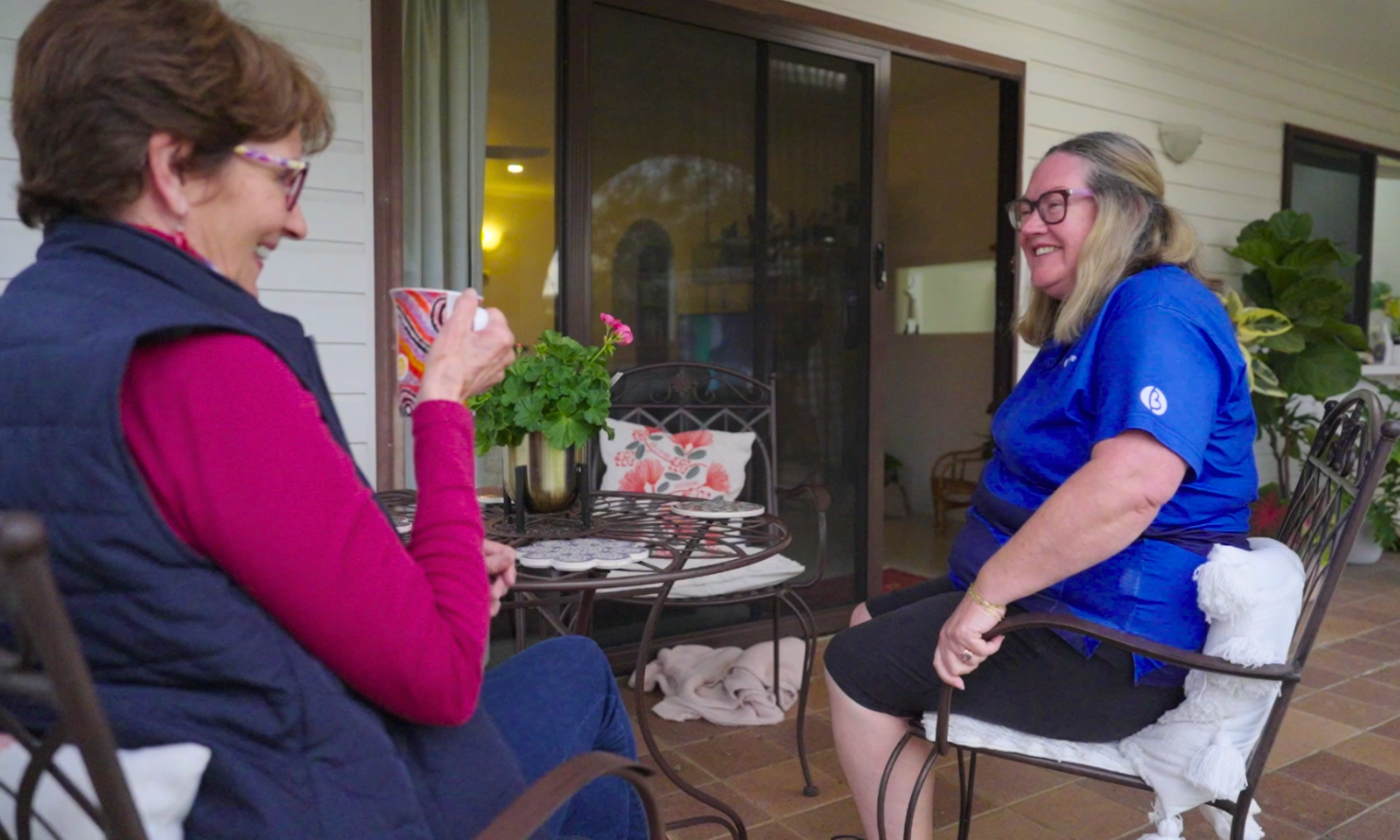Jane and Helen sitting on a patio and talking while holding drinks. Jane is wearing a pink shirt and a navy vest, and Helen is wearing her BlueCare uniform.