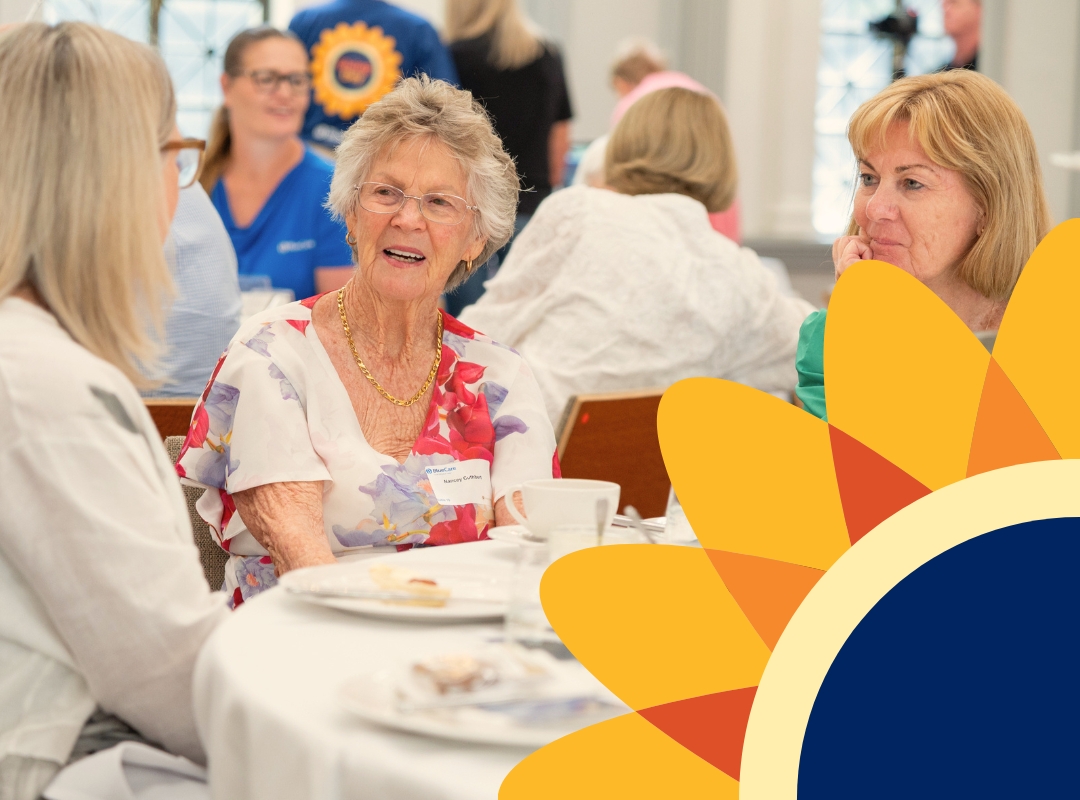 Donors seated at high tea tables indoors, chatting and eating; an elderly woman in a red floral top smiles while another woman in a green blouse listens.