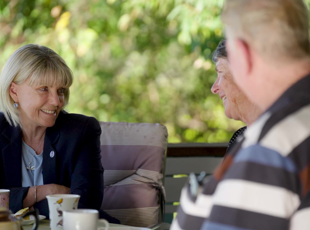 Dementia consultant and educator, Sherri, is sitting at the outdoor table of an elderl couple, smiling and talking; teacups and saucers are on the table, with soft green foliage in the background and one person in a striped sweater in the foreground.