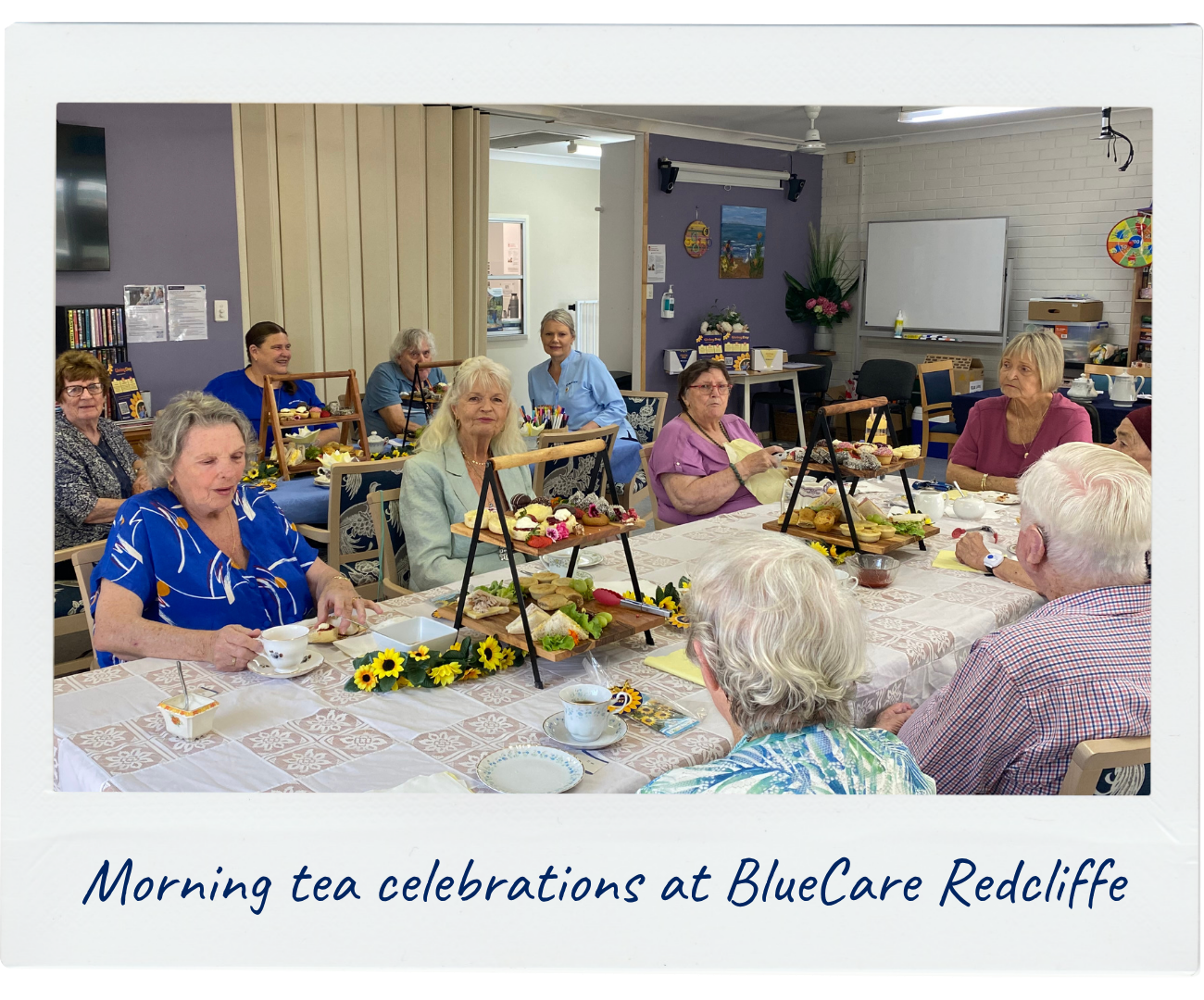 A large group of people who call BlueCare home are enjoying a delicious morning tea in a space filled with sunflowers