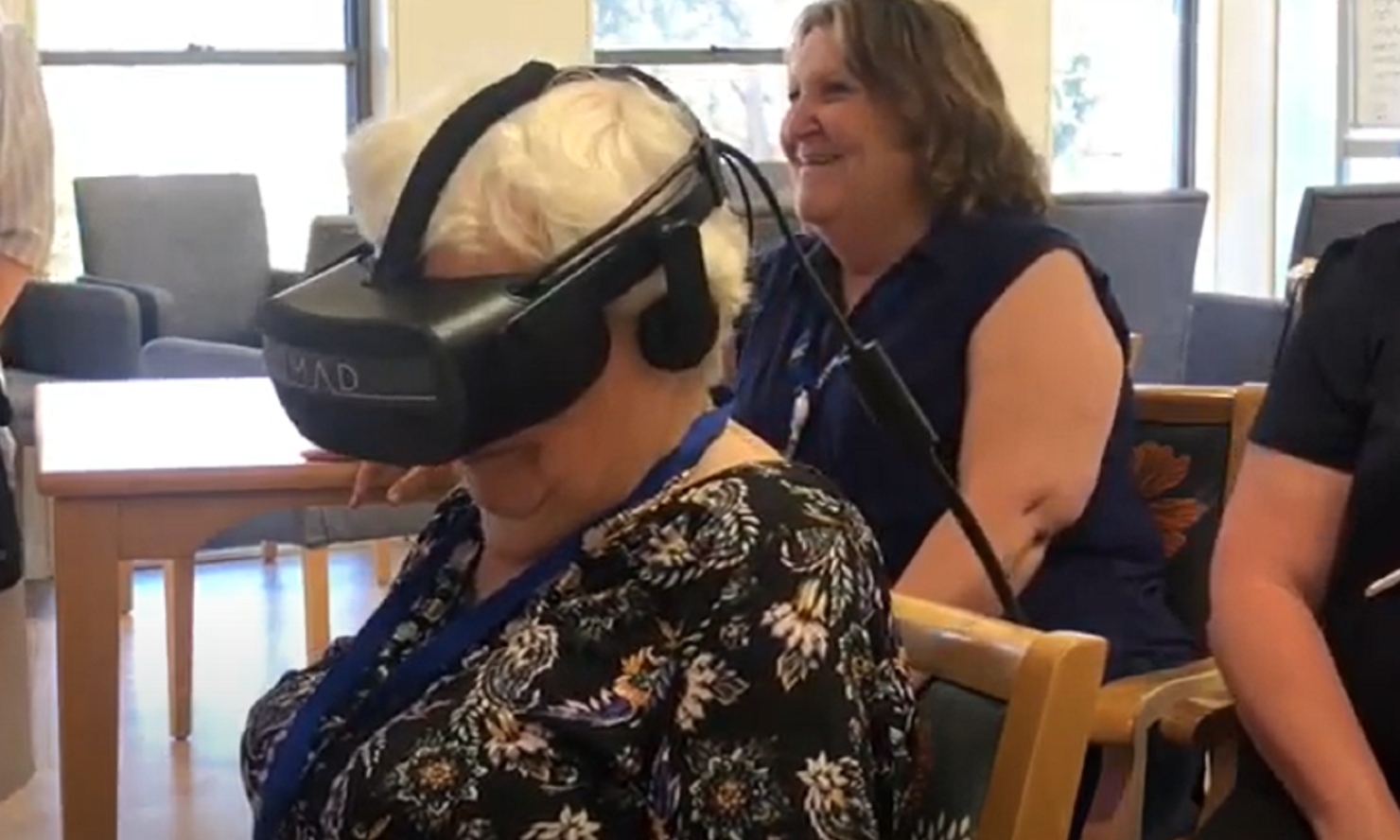 A person wearing a VR headset is seated, with another person smiling in the background.