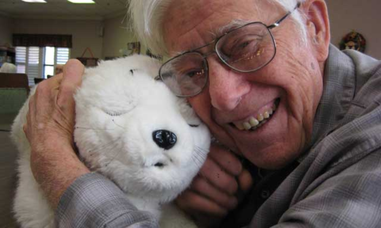 An elderly person wearing glasses smiles while hugging a white robotic pet seal.
