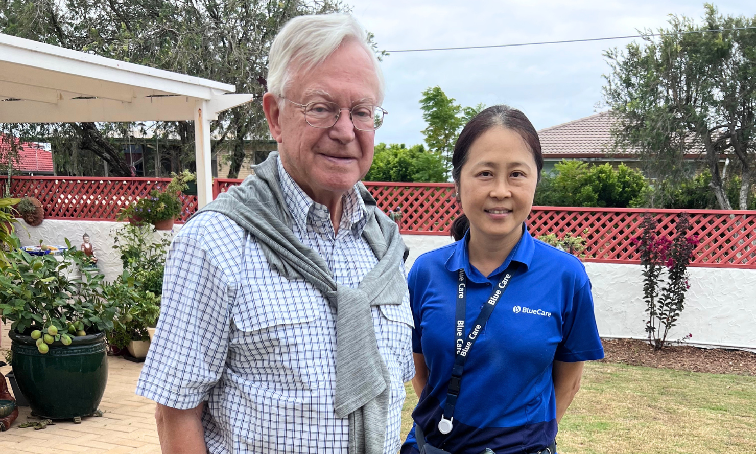 Errol and one of his specialist dementia carers, Lan, stand in Errol's garden in front of a white wall with a red lattice fence and some potted plants.