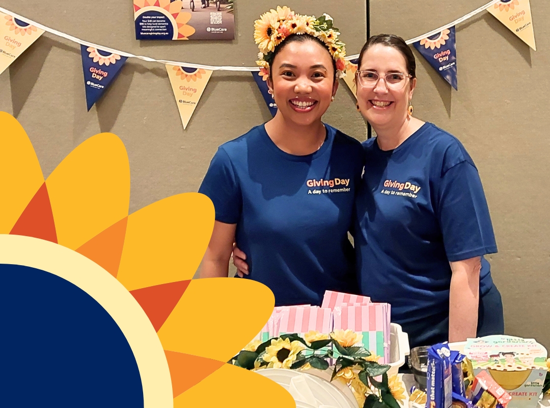 Two people wearing matching BlueCare Giving Day T-shirts stand behind a decorated table with a treats, flowers, blue party items, and bunting flags overhead at a fundraising stand