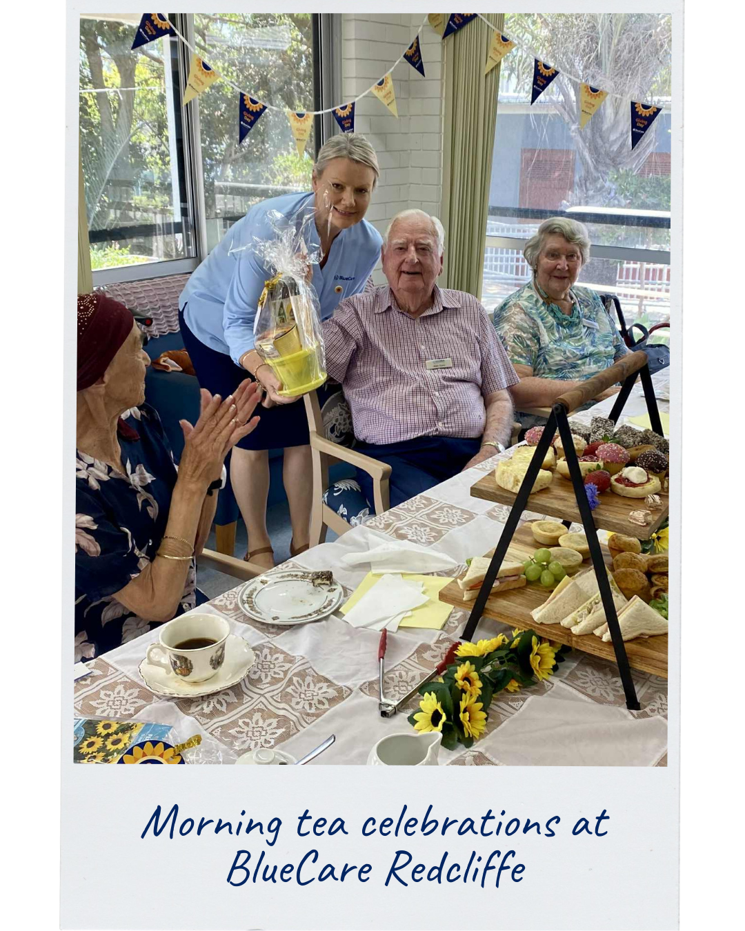 A group of people who call BlueCare home are enjoying a morning tea together. One gentleman is holding up a raffle prize he received from a BlueCare team member. The morning tea is brightly decorated with sunflowers, bunting and delicious treats