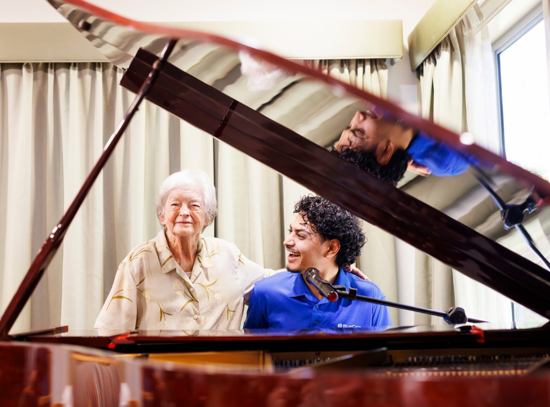 Older woman seated at a piano while a BlueCare Carer leans in beside her; the piano lid and strings frame the scene, with soft light and curtains in the background.