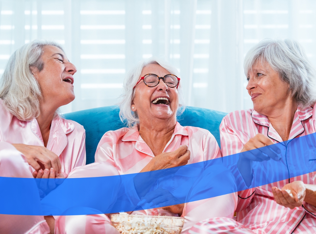 Three women in pink pajamas laugh while sitting on a blue couch with a bowl of popcorn.