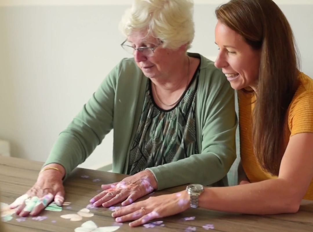 A elderly person and a young carer sit at a table playing with the light of a Magic Table, placing colorful pieces while smiling; one person in a green top uses hands to arrange tiles, with hands and cards visible.
