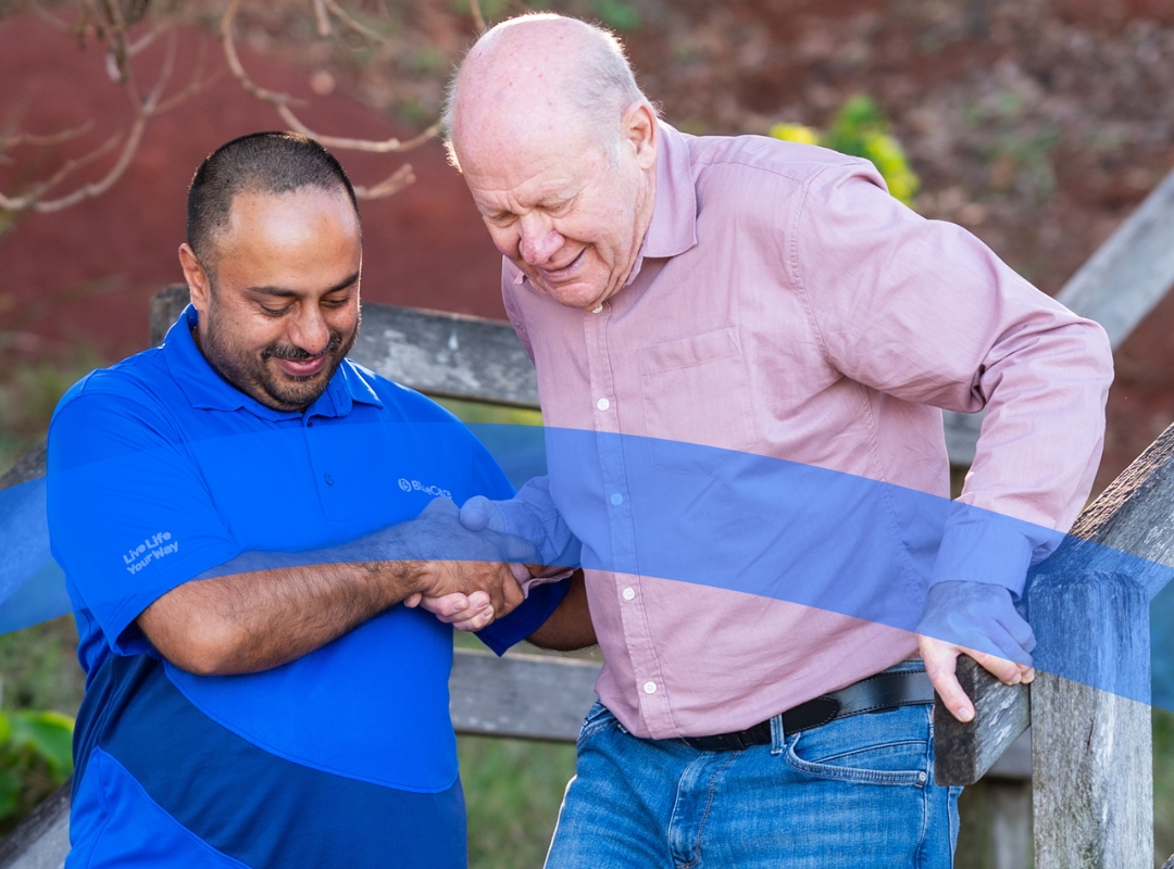 A BlueCare Personal Carer in a blue shirt holds the hand of an elderly man in a pink shirt while helping him down a wooden staircase to the beach.