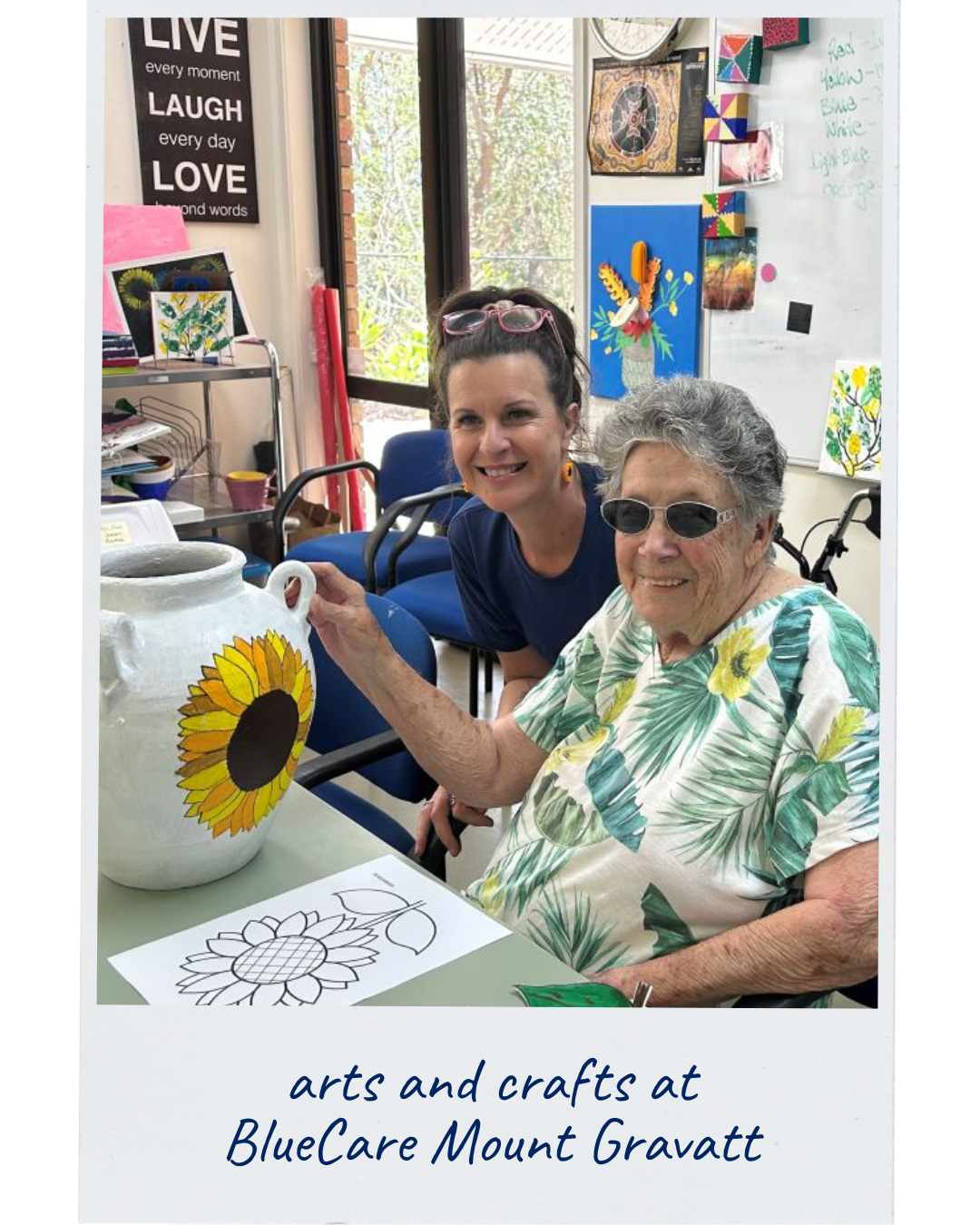 An older woman and a BlueCare team member are decorating a large vase with a vibrant sunflower design
