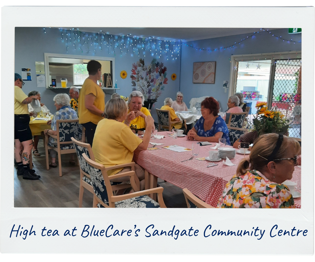 A group of older people are enjoying a delicious high tea at their local BlueCare Community Centre. There are sparkling blue lights and lots of bright yellow shirts filling the space.