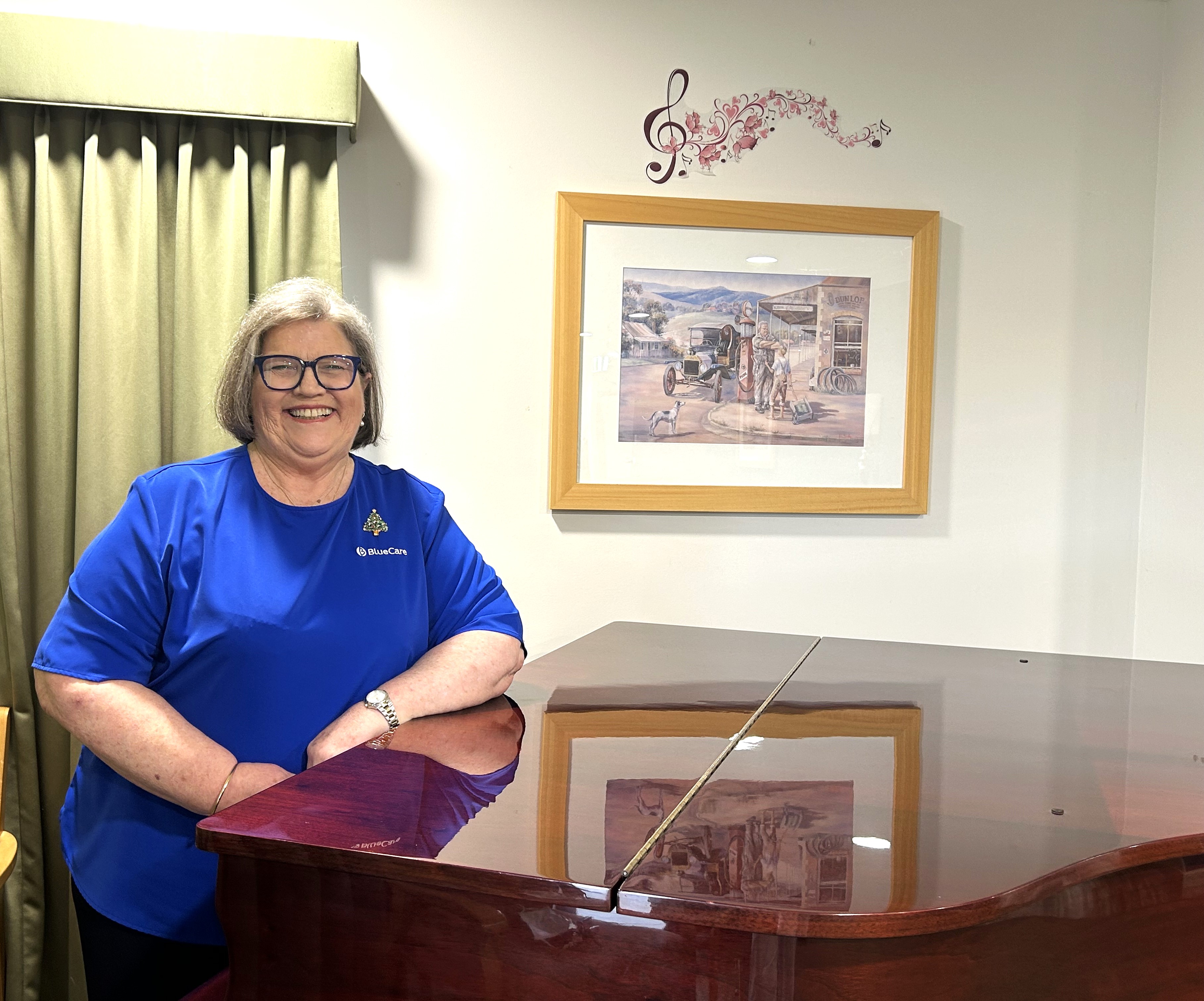 A photo of Catherine Sherlock. Cathering is leaning against a piano in the community centre of her local BlueCare Aged Care Home. Catherine has a bright blue shirt, matching glassess and an infectious smile.