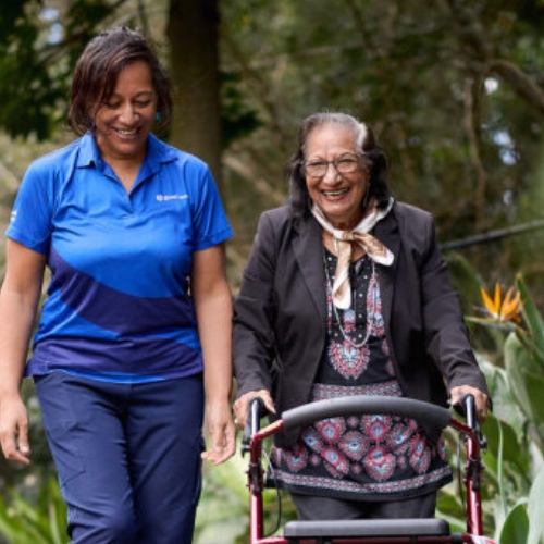A BlueCare team member and an older woman who calls BlueCare home are out for a stroll together in beautiful green parklands