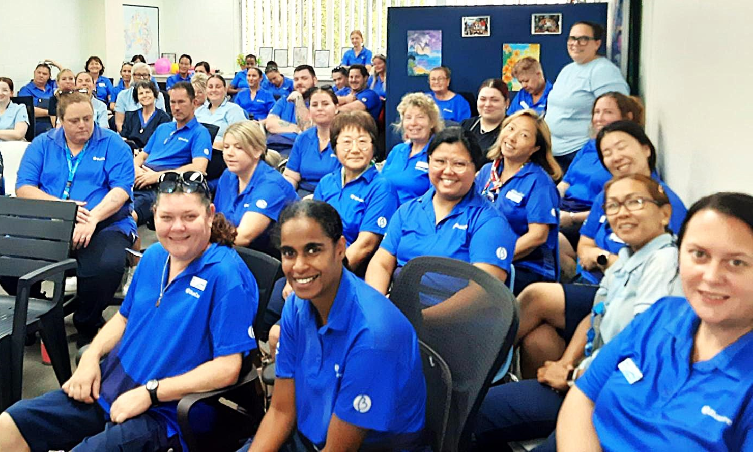A diverse group of BlueCare carers and nurses wearing matching BlueCare blue shirts are seated in a room training room, looking towards the camera.