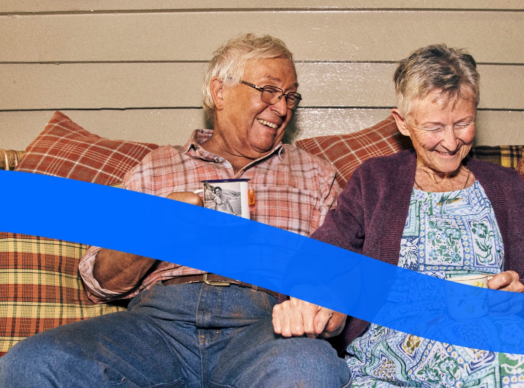 An elderly couple sits on a couch holding mugs and holding hands, smiling at each other.